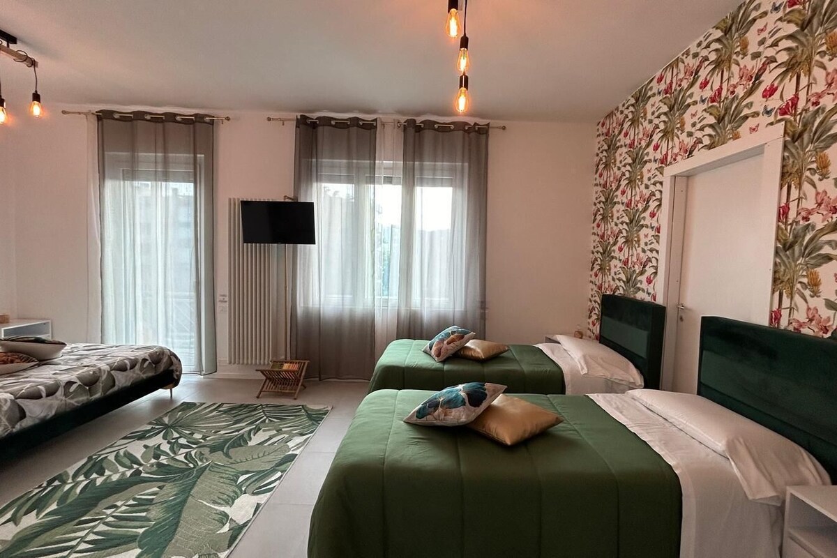 Una camera d'albergo con due letti, una TV a schermo piatto, carta da parati con motivo floreale e una finestra con tende trasparenti.