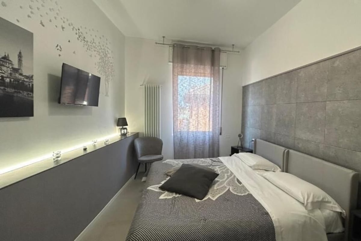 Una camera d'albergo moderna con un grande letto, una TV a schermo piatto fissata al muro, un comodino con una lampada e una finestra con tende.