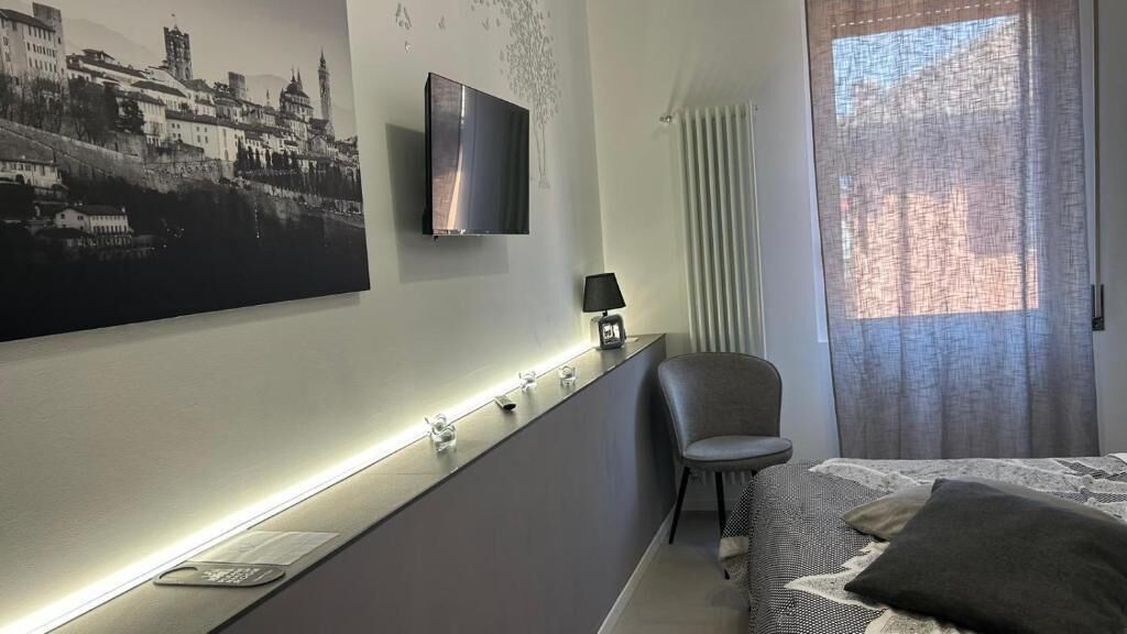 A modern hotel room with a bed, a chair, a TV mounted on the wall, and a cityscape painting.
