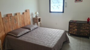 A bedroom with a wooden headboard, a bed with a patterned bedspread, a nightstand with a lamp, a dresser with drawers, and a window with a leaf pattern. - Gîte "la Galinette" in village center, air-conditioned with swimming pool. (Vallon-Pont-d'Arc)