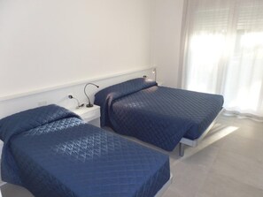 Two single beds with blue quilts, a white nightstand with a lamp, and a window with sheer curtains. - Cozy Flat for Five Guests in an Enclosed Residence (San Michele al Tagliamento)