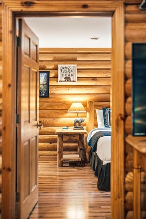 Ein Interieur einer Holzhütte mit einem Bett, einem Nachttisch mit Lampe und einem gerahmten Schild mit der Aufschrift 'DON'T WOKE THE BEAR'.