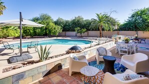 Ein Pool mit Liegestühlen und Tischen, umgeben von Grünflächen und blauem Himmel.