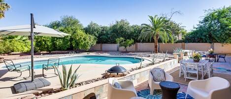 Une piscine entourée de verdure, avec des chaises longues et des tables, sous un ciel dégagé.