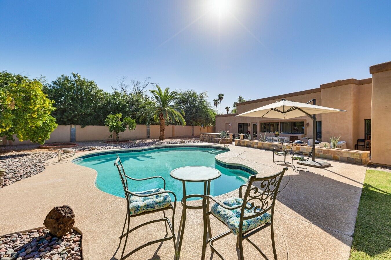 A swimming pool with two chairs and a table, surrounded by a patio area with a building and trees.