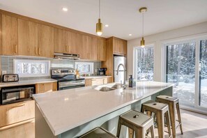 A modern kitchen with wooden cabinets, stainless steel appliances, a large island with stools, and a view of a snowy landscape through large windows. - Private Pool & Spa | Chalet Bella Vista (Saint-Côme)