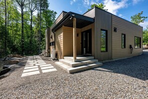 A modern building with a covered porch, a gravel driveway, and a forested background. - Private Pool & Spa | Chalet Bella Vista (Saint-Côme)