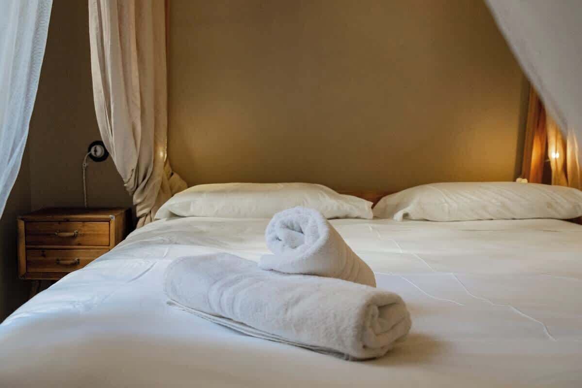 A neatly made bed with white linens and a rolled towel, a wooden nightstand with drawers, and a wall clock.