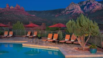 A pool area with lounge chairs and umbrellas, surrounded by a fence, with mountains in the background.