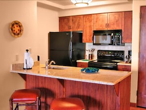 A kitchen with wooden cabinets, a black refrigerator, a microwave, a stove, and a bar area with stools. - Hyatt Pinon Pointe (Sedona)