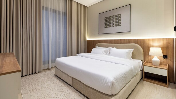 Quarto de hotel com uma cama grande, uma mesa de cabeceira com luminária, um quadro na parede e uma janela com cortinas.