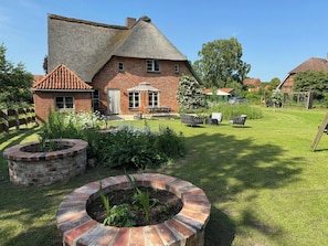 Ein Backsteinhaus mit Strohdach, ein Garten mit kreisförmigem Ziegelpflanztopf und eine Terrasse mit Gartenmöbeln.