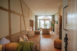 Ein modernes Wohnzimmer mit Holzbalkendecke, einer Couch mit gemusterten Kissen, einem Esstisch und einem großen Fenster.