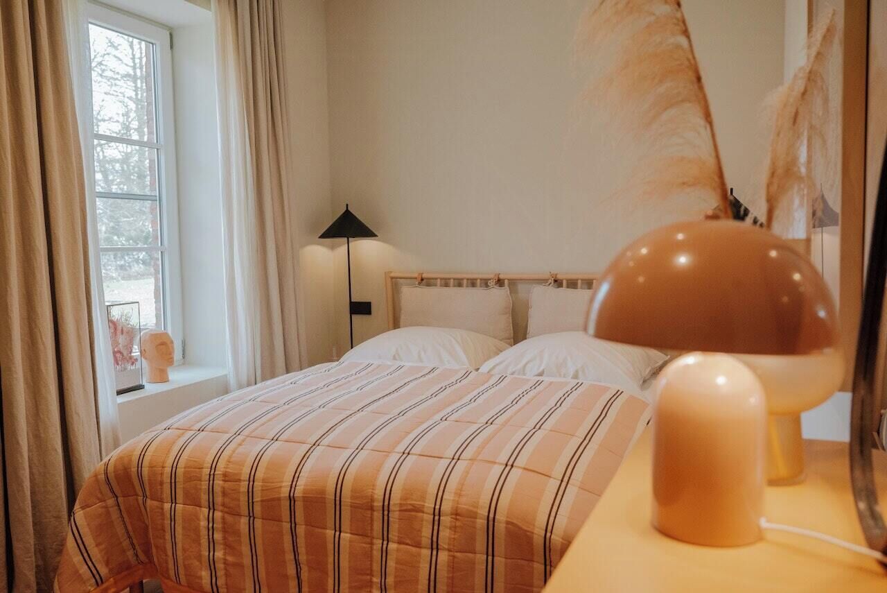 Een slaapkamer met een bed, een bedlampje, een raam met gordijnen en een spiegel aan de muur.