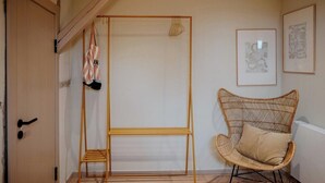 Ein Zimmer mit einer Holztür, einem Rattanstuhl, einem Metallständer und gerahmten Kunstwerken an der Wand.