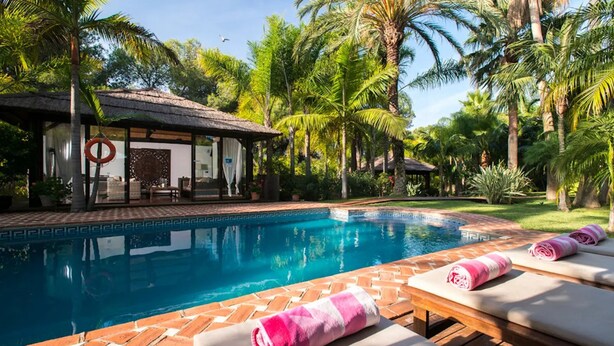 A swimming pool with lounge chairs and a covered seating area, surrounded by palm trees and greenery. - Villa in palm tree plantation, near the beach. (Marbella)