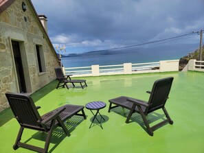 Eine Terrasse mit zwei Stühlen und einem Tisch, mit Blick auf ein Gewässer und ein Steinhaus.