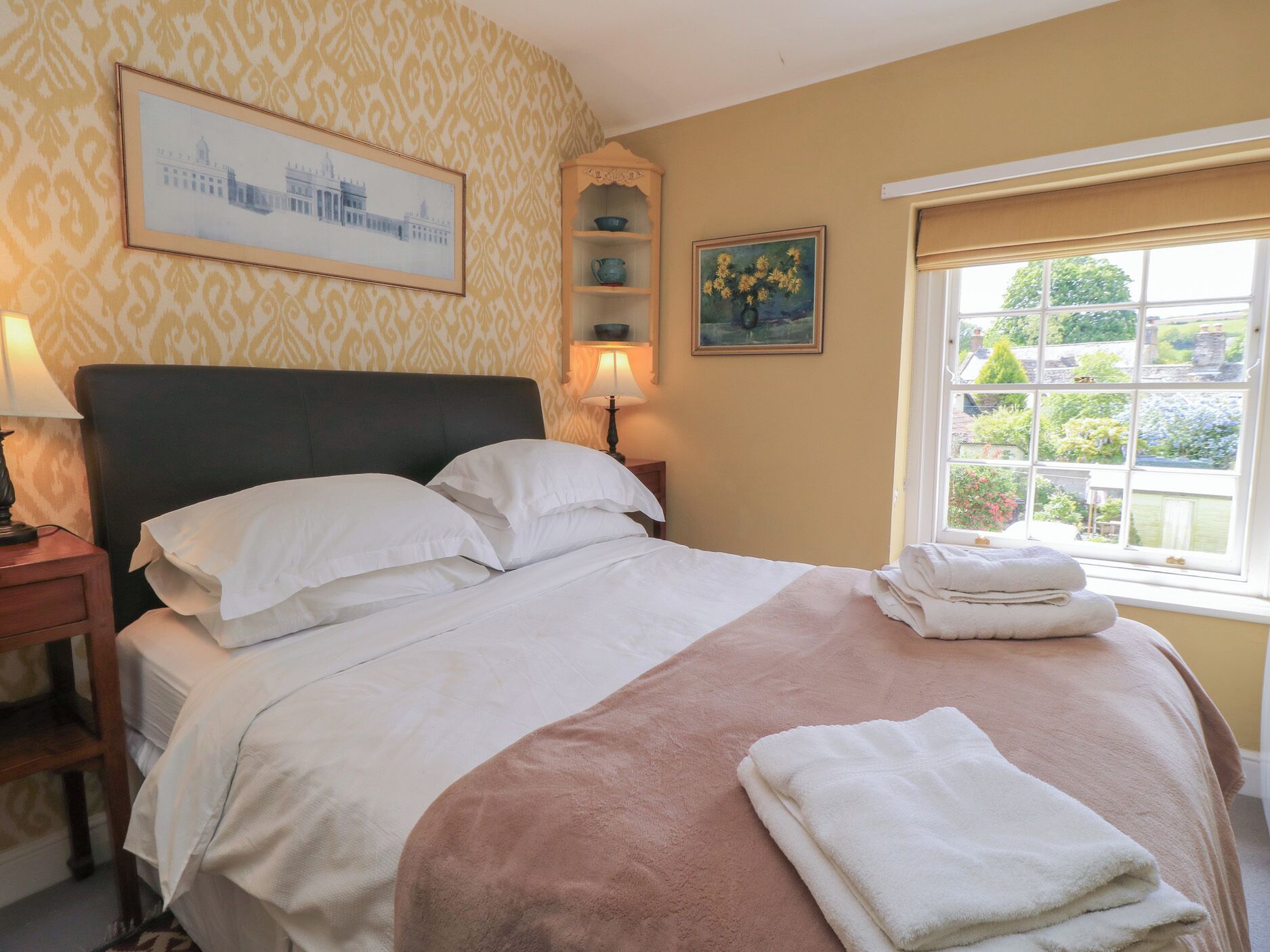 A neatly made bed with white and pink linens, a framed picture of a building on the wall, and a window with a view of greenery.