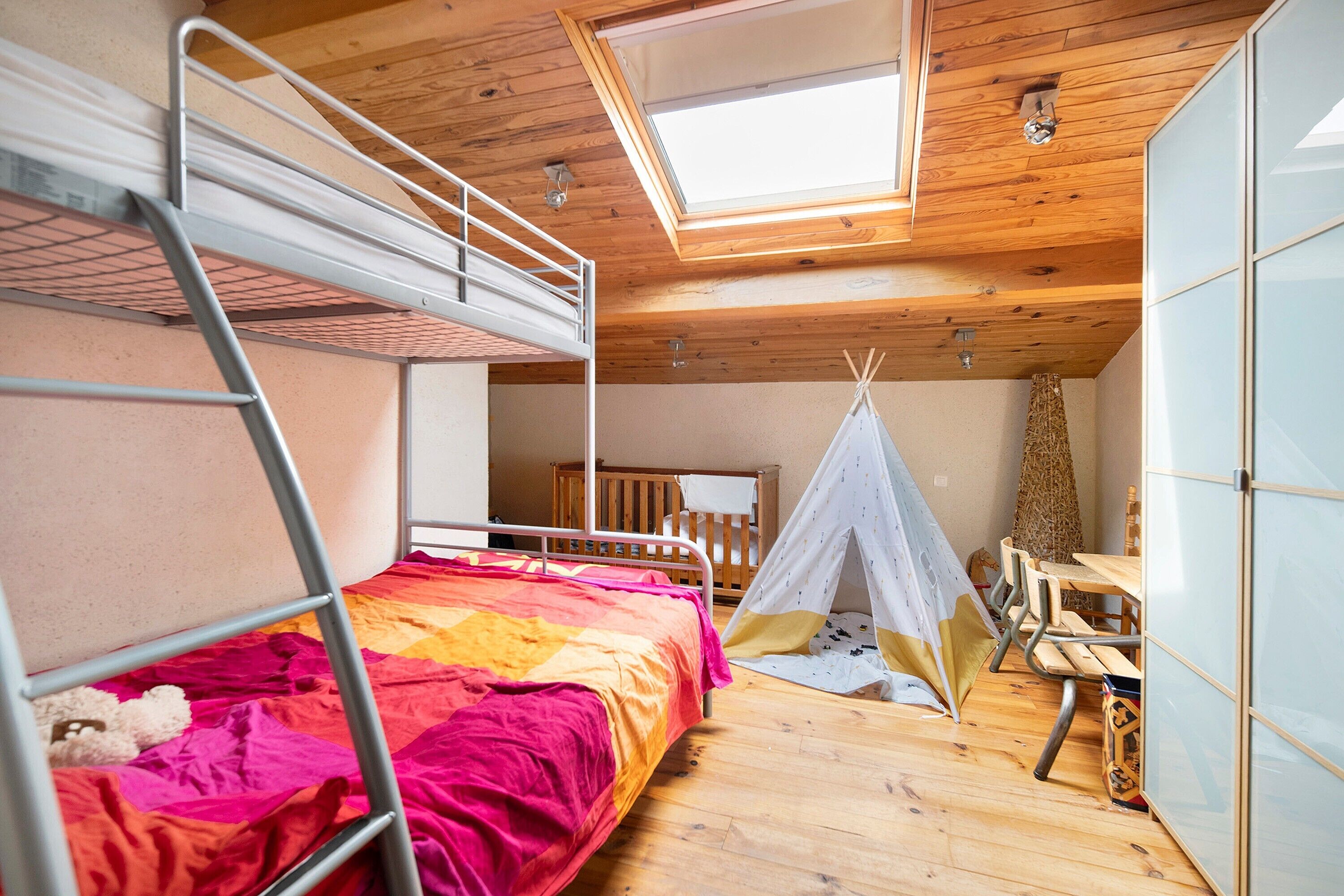 Une pièce avec des lits superposés, un plafond en bois, une lucarne, une petite tente et une table avec des chaises.