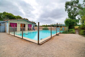 Une piscine entourée d’une clôture, un petit bâtiment avec un toit rouge et un chemin de gravier.