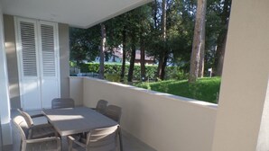 A balcony with a table and chairs, a white door with shutters, and a view of a green landscape with trees. - New Flat for Five Guests in a Residence With Pool (San Michele al Tagliamento)
