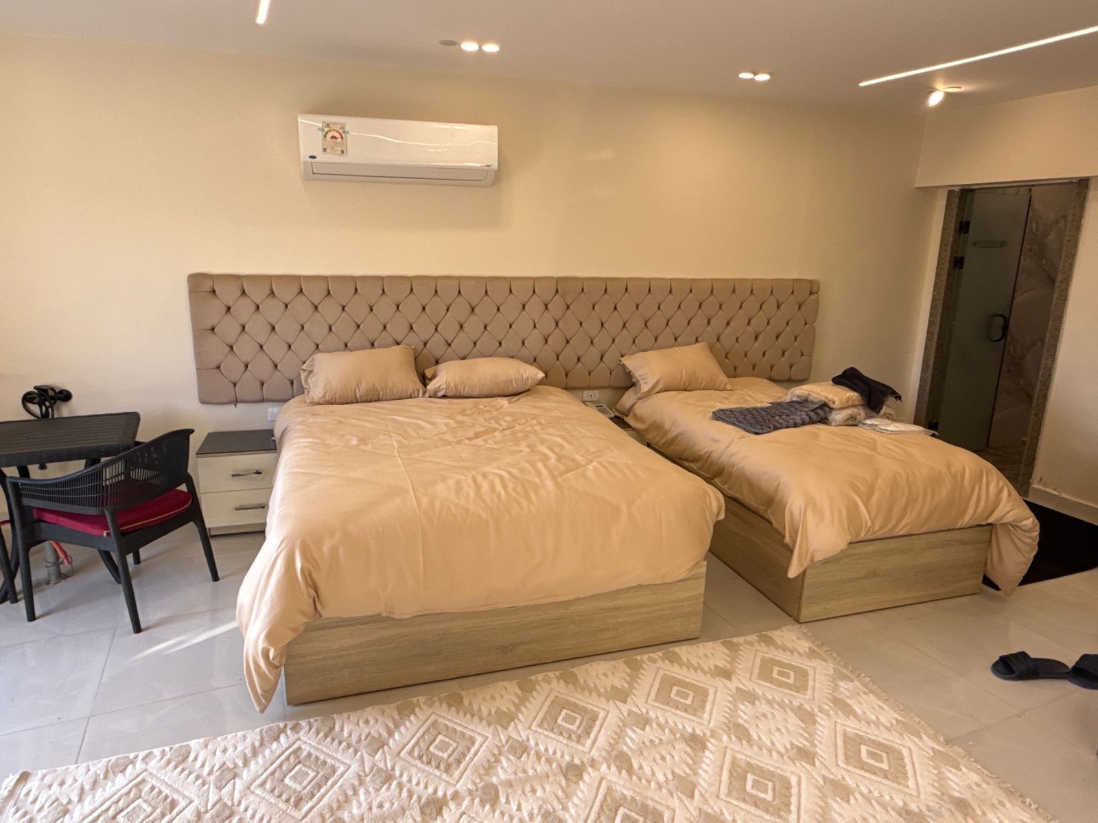 A bedroom with two beds, a desk, a chair, an air conditioning unit, and a patterned rug.