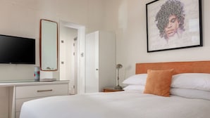 Una habitación de hotel con una cama, un escritorio con televisión de pantalla plana, un espejo, una obra de arte enmarcada y una mesita de noche con lámpara.