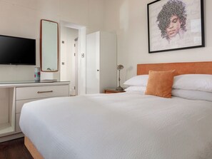 Ein Hotelzimmer mit Bett, Schreibtisch, Flachbildfernseher, Spiegel, einem gerahmten Kunstwerk und einem Nachttisch mit Lampe.