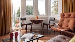 Una habitación con ventanal, un sofá, dos sillones, una mesa con un libro y tazas, y vistas a la vegetación exterior.