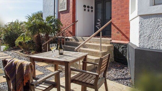 Un área de estar al aire libre con madera, que incluye una mesa, sillas, una botella de vino y un letrero que dice 'Chelsea House'.