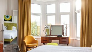 Quarto de hotel com cama, escrivaninha com espelho, cadeira e janela com cortinas.