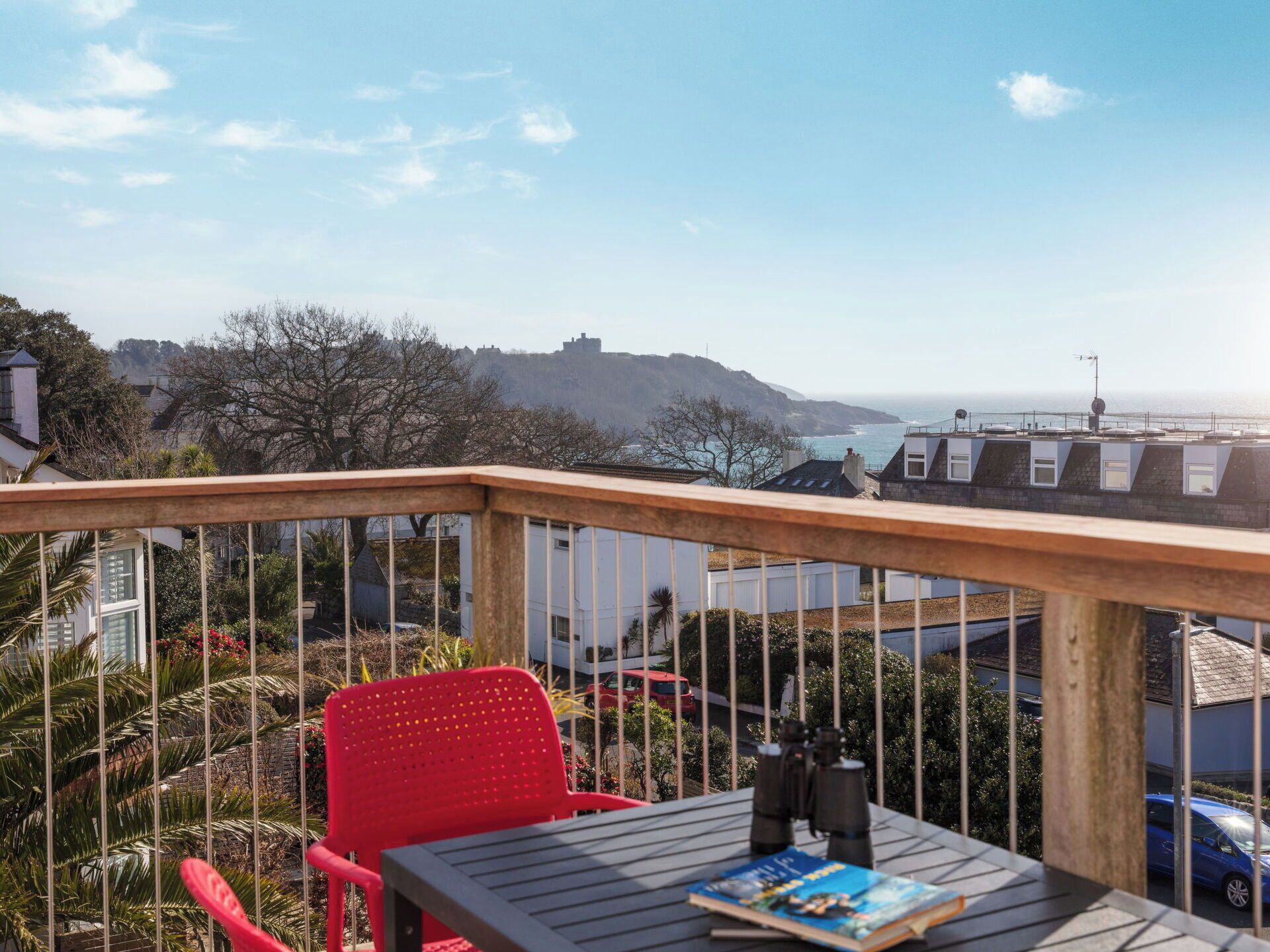 A balcony with a red chair, a table with binoculars and a book, and a view of the sea and a hill in the background.