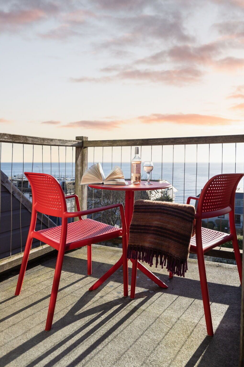Un balcon avec des chaises rouges, une table, une bouteille de vin et un livre ouvert.