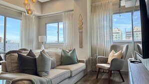 Una sala de estar moderna con sofá, sillones, una mesa de centro y un mueble para la televisión. Ventanas amplias ofrecen vistas del paisaje urbano.