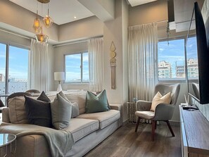 Una sala de estar moderna con sofá, sillones, una mesa de centro y un mueble para la televisión. Ventanas amplias ofrecen vistas del paisaje urbano.