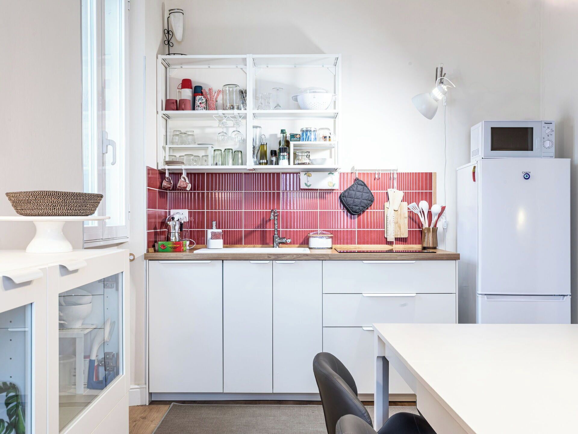 A modern kitchen with white cabinets, a red-tiled backsplash, a white refrigerator, and a dining area with a table and chairs.