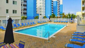 A swimming pool with blue lounge chairs and a black umbrella, surrounded by a brick patio and a multi-story building with balconies. - Purnell House 206 (Ocean City)