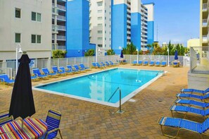 A swimming pool with blue lounge chairs and a black umbrella, surrounded by a brick patio and a multi-story building with balconies. - Purnell House 206 (Ocean City)