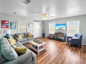 Ein Wohnzimmer mit grauem Sofa, einem Couchtisch, einem blauen Sessel, einem Deckenventilator und einem Fernseher, der eine Strandansicht zeigt.