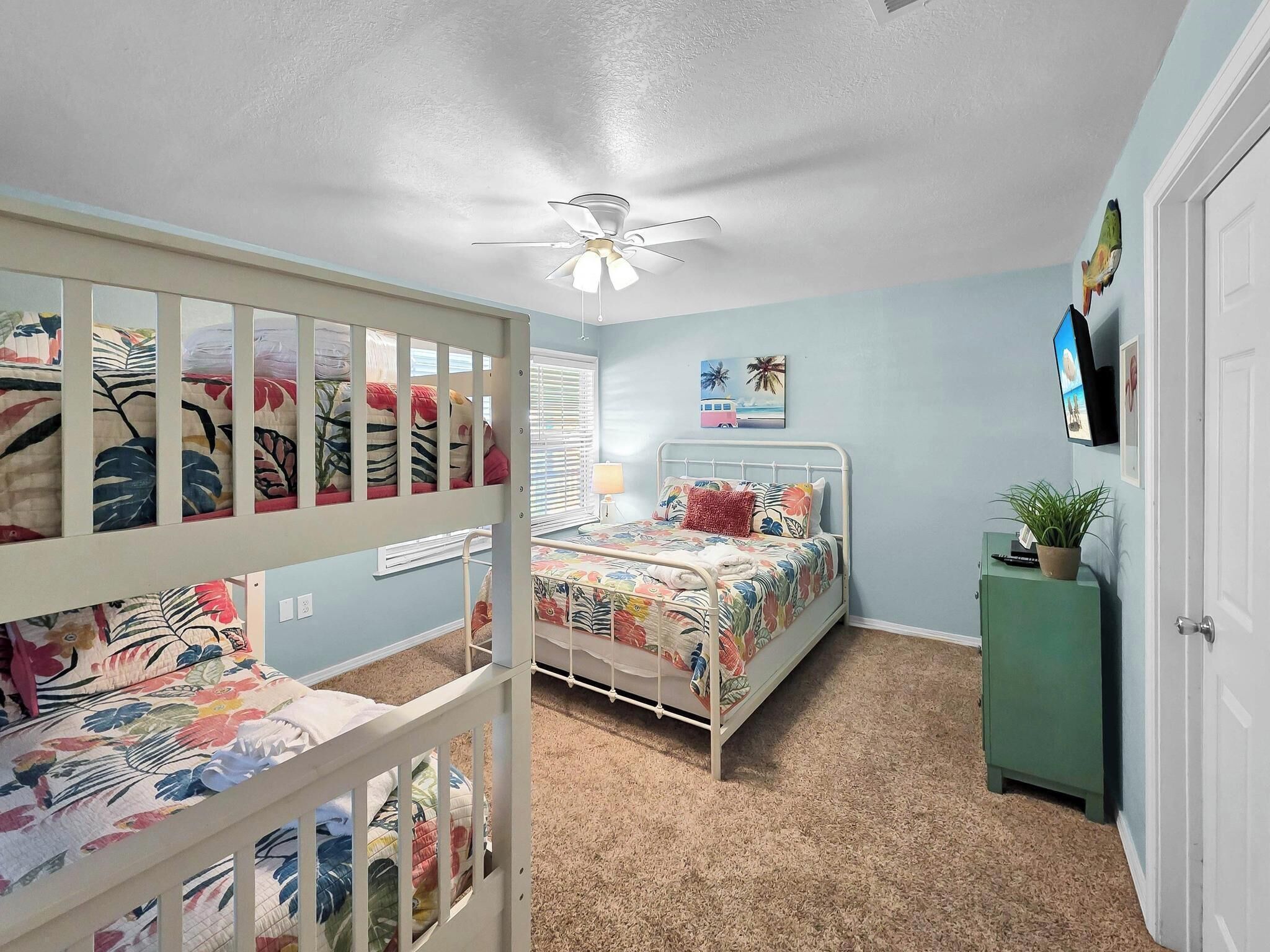 A child's room with bunk beds, a ceiling fan, a bed with floral bedding, a green cabinet, and a wall-mounted TV.