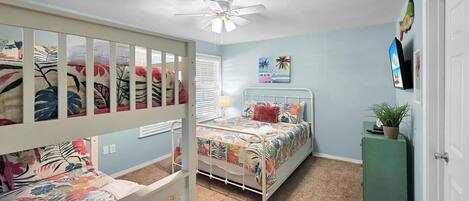A child's room with bunk beds, a ceiling fan, a bed with floral bedding, a green cabinet, and a wall-mounted TV.