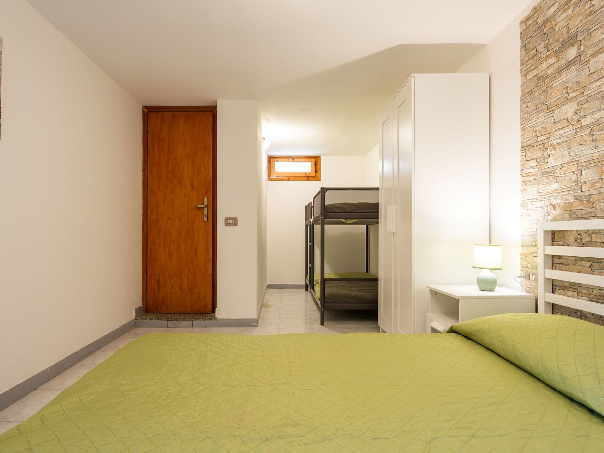 A bedroom with a bunk bed, a wooden door, a stone wall, and a bedside lamp.