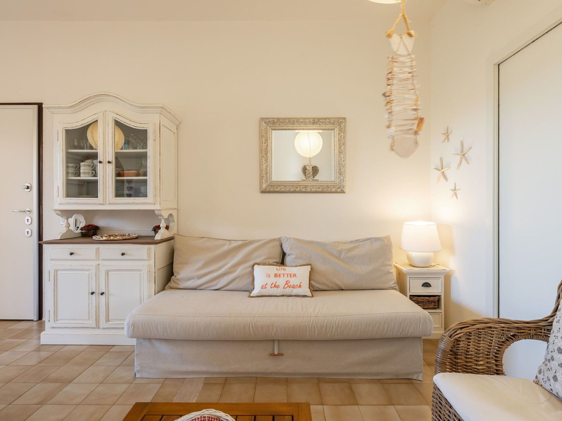 Una habitación acogedora con un sofá, un aparador con puertas de cristal, un espejo enmarco, una mesita de noche con lámpara y una silla de mimbre.