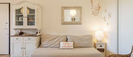 Una habitación acogedora con un sofá, un aparador con puertas de cristal, un espejo enmarco, una mesita de noche con lámpara y una silla de mimbre.
