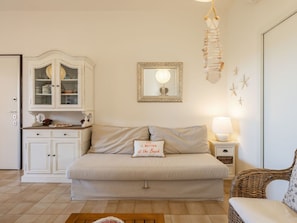 Ein gemütliches Zimmer mit einer Couch, einem Schrank mit Glastüren, einem gerahmten Spiegel, einem Nachttisch mit Lampe und einem Korbstuhl.