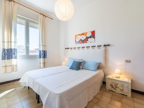 Ein Schlafzimmer mit Bett, Vorhängen, einem Nachttisch, einer Lampe, einem Wandradiator und einem Fenster.