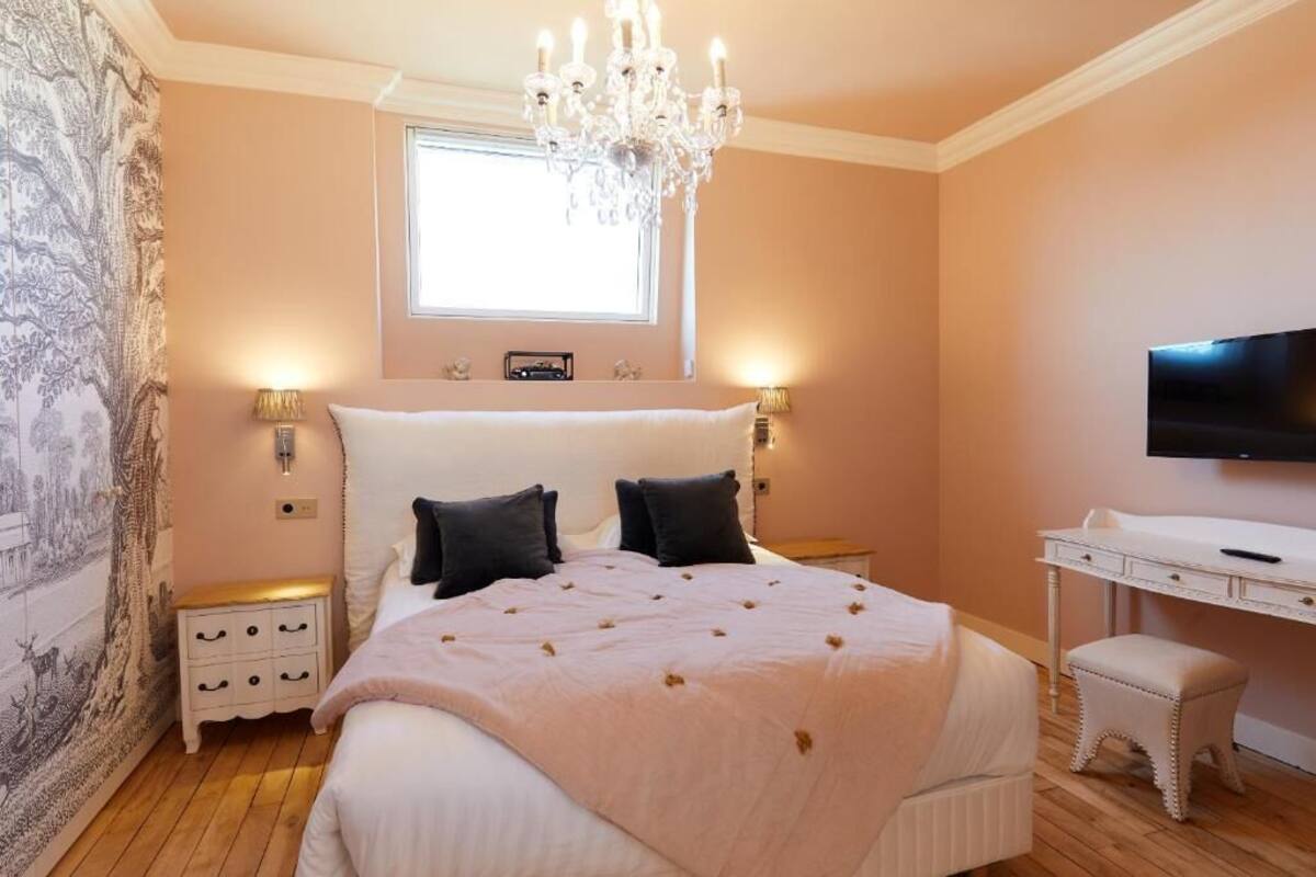 A bedroom with a bed, a chandelier, a TV, a desk, and a window.