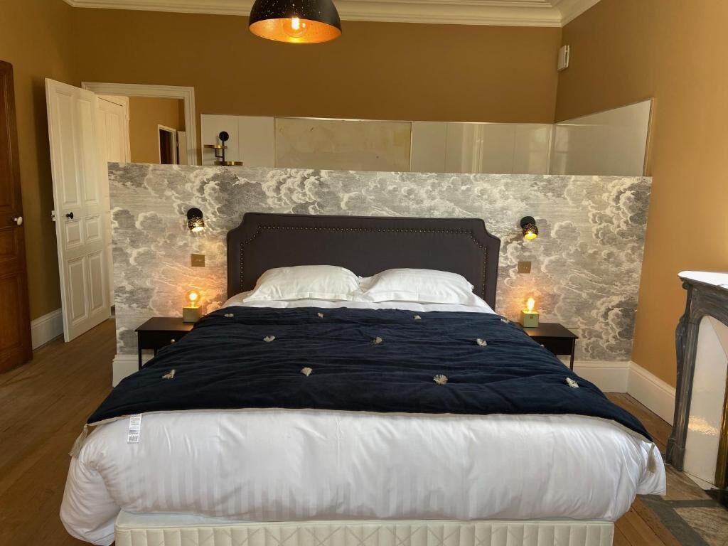 Un dormitorio con cama, mesitas de noche, espejo y lámpara de pared.