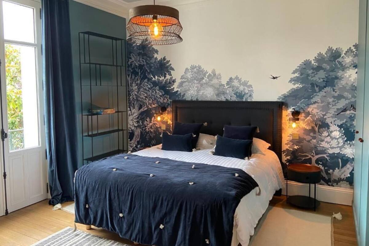 A bedroom with a bed, a hanging light fixture, a wall mural, and a window with curtains.