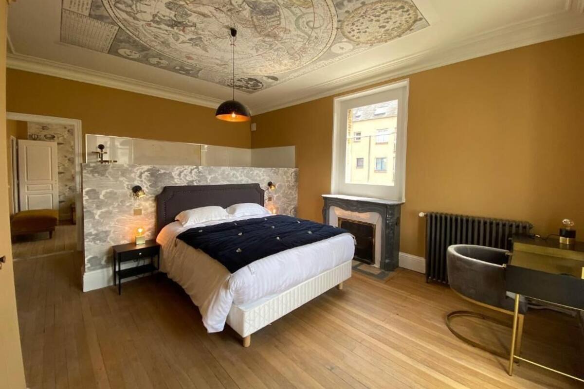 A bedroom with a bed, nightstands, a fireplace, a desk, and a map-tiled ceiling.